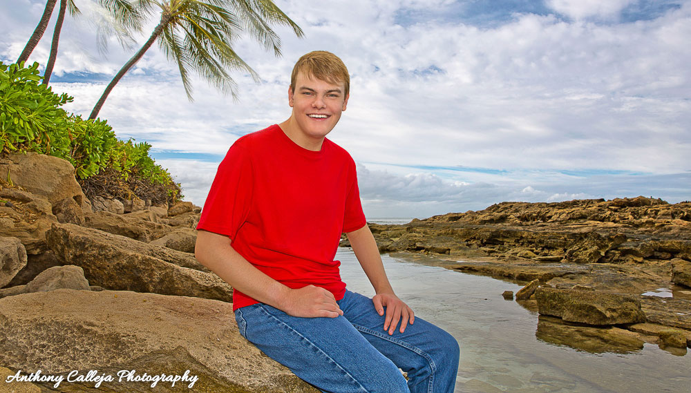 Portrait of Oahu Graduating Senior, sitting on a Rock at  Paradise Cove Beach, Oahu. Beautiful seascape and palms trees in the background.